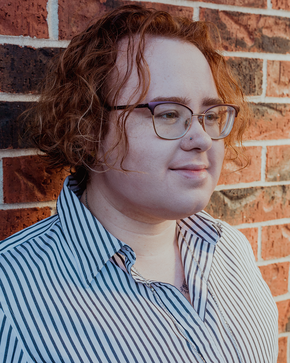 A headshot of Magic. They are facing to the right at a 3/4 view wearing a collared button up with vertical black and white stripes. The top two buttons are undone. They are wearing large glasses that are a dark purple and their nose stud is visible. Behind them is a red brick wall. 